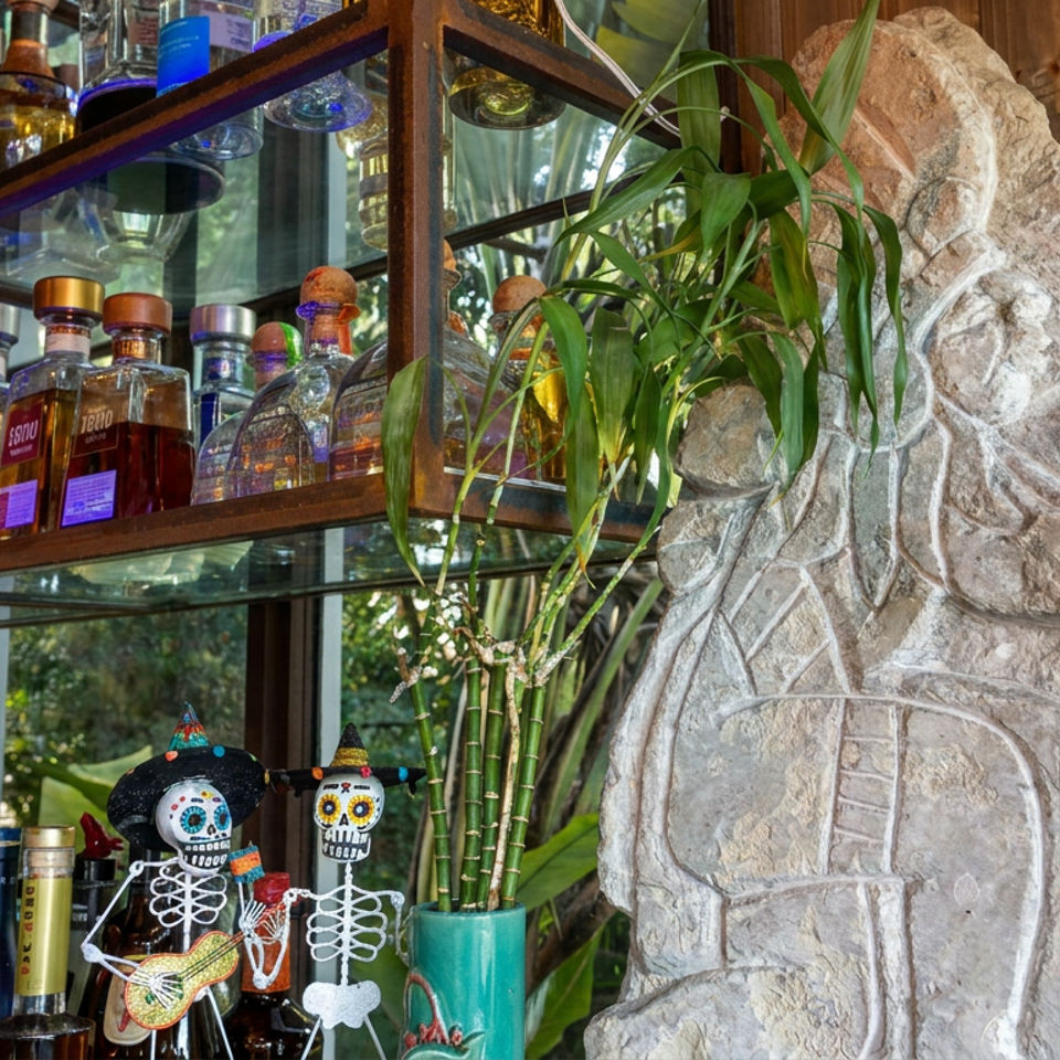 Restaurant bar displaying tequila bottles, glassware, bamboo plants, and colorful Día de los Muertos skeleton decorations beside a carved stone sculpture.